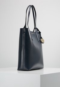 Marineblauer Ledertote mit zwei langen Griffen, flachem Boden, strukturierter Oberfläche und einem goldfarbenen Akzentanhänger. Einfaches rechteckiges Design.