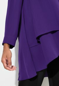 Fialová, volná blůza s dlouhými rukávy, asymetrickým spodním lemem a jemnou texturou. Obsahuje vrstvený design a široké detaily manžet.