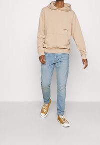 Sudadera con capucha beige con bolsillo delantero y cordones en la capucha, combinada con jeans azul claro y zapatillas amarillas. Sobre un fondo liso.