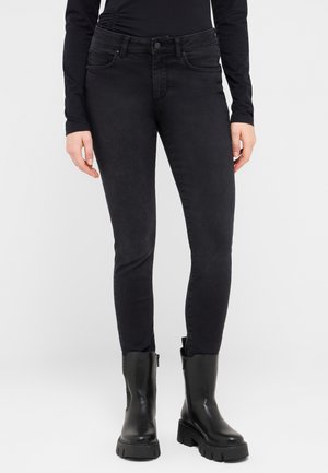 Juodos spalvos siauri džinsai, turintys aukštą liemenį, subtilų išblukimą ir priekinės kišenės, derinami su juodais odiniais aulinukais iki kelių.