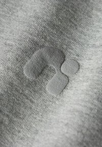 Un tessuto grigio con una sottile texture presenta un logo in rilievo e liscio di una tonalità corrispondente, enfatizzando un design minimalista.