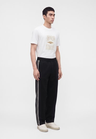 Νέος άνδρας που φορεί λευκό T-shirt Emporio Armani, μαύρο παντελόνι με λευκές ρίγες στα πλάγια και λευκά αθλητικά παπούτσια.