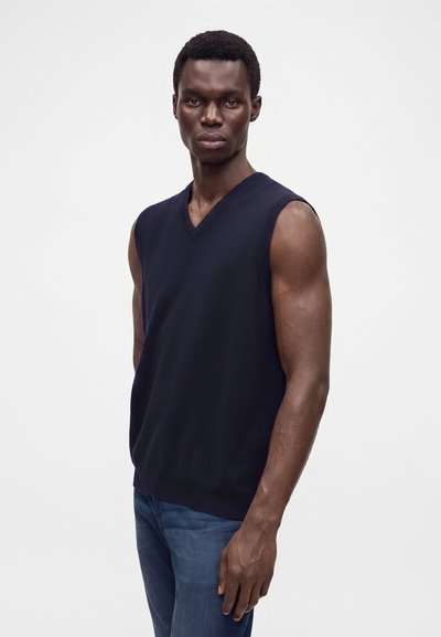 Homme portant un pull sans manches bleu marine foncé à col en V et un jean bleu, debout devant un fond blanc uni.