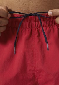 Pantaloni da bagno rossi con vita elasticizzata, cordoncino navy e piccolo etichetta con logo. Texture liscia con un leggero lucido. Primo piano dell'area vita.