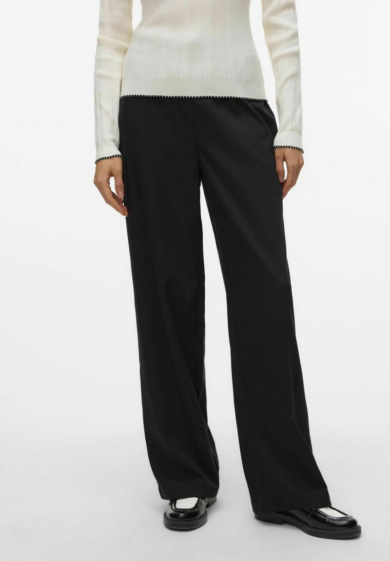 Pantalon large noir en tissu lisse, offrant une coupe décontractée et des plis avant discrets. Associé à un haut blanc à côtes.