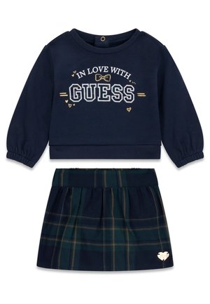 Haut à manches longues bleu marine avec le texte « In Love With Guess » associé à une jupe à carreaux bleu marine et vert foncé avec un emblème en forme de cœur.