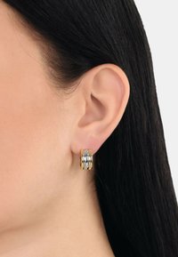 Orecchini a cerchio a tre tonalità con anelli intrecciati in finiture argentate, dorate e rosé, esposti su un orecchio contro capelli scuri.