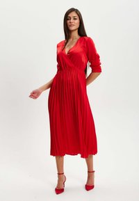Red pleated dress with a deep V-neckline and long sleeves, made from a smooth fabric. Worn with matching red high-heeled shoes.