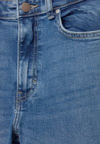 Krupan prikaz plavih traperica s metalnom kopčom, petljom za pojas, prednjim džepom i detaljnim šavovima.
