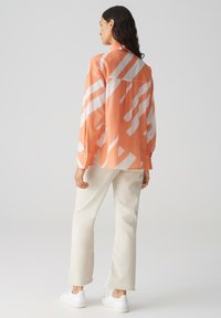 Blusa arancione con motivi geometrici bianchi, maniche lunghe, polsini con bottoni, tessuto leggero, abbinata a pantaloni bianchi e sneakers.