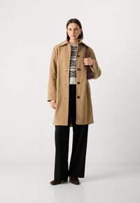 Manteau en laine beige avec un col, des boutons sur le devant et des fentes sur les côtés. Porté par-dessus un pull en maille avec des motifs noir et blanc, assorti à un pantalon noir.