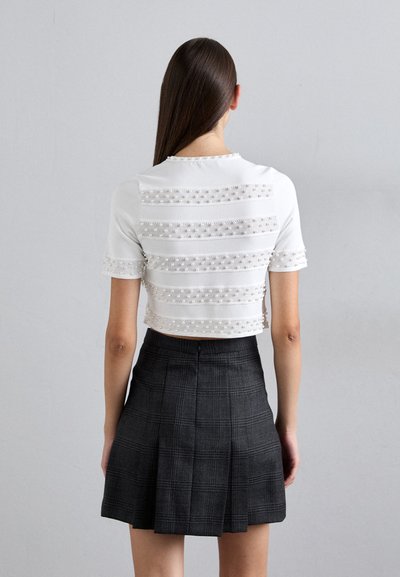 Une femme aux longs cheveux bruns porte un crop top blanc avec des embellissements en perles et une jupe plissée taille haute grise foncée, se tenant contre un mur gris.