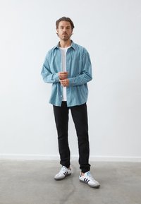 Hombre de pie en interiores que lleva una camisa a rayas azul y blanca sobre una camiseta blanca, pantalones negros y zapatillas blancas con rayas negras.