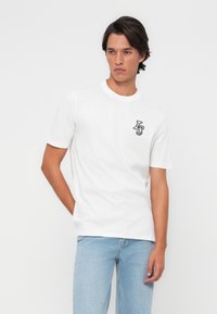Hvit bomull t-skjorte med korte ermer, rund halslinning og svart brodert "LS"-logo på venstre bryst. Brukt med lyseblå jeans.
