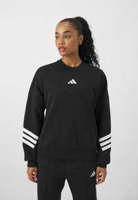 Sweatshirt noir avec un col rond, coupe surdimensionnée et motif à trois rayures blanches sur les manches. Logo minimaliste sur la poitrine.