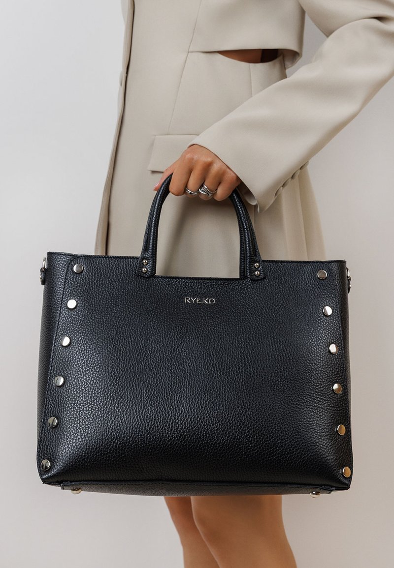 Person in beige coat holding black textured leather handbag with silver studs and "RYŁKO" logo in gold lettering.