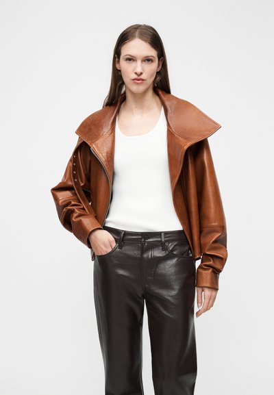 Γυναίκα που φοράει καφέ δερμάτινο μπουφάν πάνω από λευκή αμάνικη μπλούζα και μαύρο δερμάτινο παντελόνι, ποζάροντας με το ένα χέρι στην τσέπη της.
