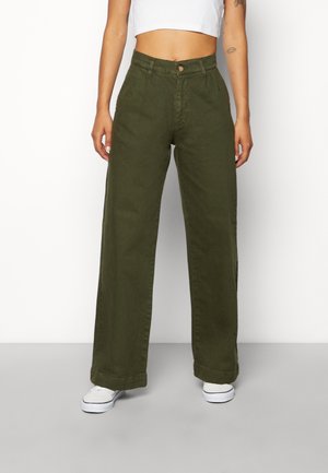 Person trägt hoch taillierte, weite olivgrüne Hose, weißes Croptop und weiße Sneakers und steht vor einem einfarbigen Hintergrund.