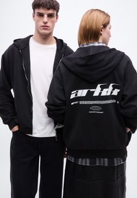 Νεαρός άνδρας που κοιτάζει μπροστά με μαύρο φούτερ με φερμουάρ και λευκό πουκάμισο· νεαρή γυναίκα με την πλάτη γυρισμένη φορώντας μαύρο φούτερ με λευκό κείμενο «arkk».