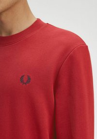 Felpa rossa con collo a giro, caratterizzata da un maglione a trama e un piccolo emblema logo blu navy sul lato sinistro del petto. Tessuto morbido e cuciture lisce.