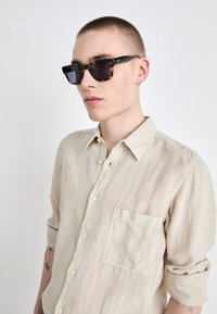 Occhiali da sole tartarugati con lenti scure, stanghette nere e bordi arrotondati. La persona indossa una camicia button-up in lino color beige chiaro con una tasca.