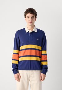 Marineblå langermet polo skjorte med kremfarget krage og horisontale striper i gult, oransje og blått. Har en liten logo på brystet.