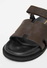 Sandal in pelle marrone con design perforato, punta aperta e cinturino posteriore. Presenta una suola nera liscia con dettaglio di cuciture sui bordi.