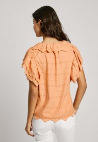 Blusa color pesca con scollo e maniche arricciati, caratterizzata da motivi floreali ad occhio di pernice e orlo ondulato, realizzata in tessuto morbido.
