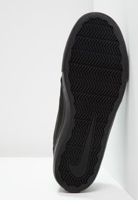 Baskets noires avec une semelle en caoutchouc texturée, présentant un motif en chevrons et un détail de logo incurvé notable sur le côté.