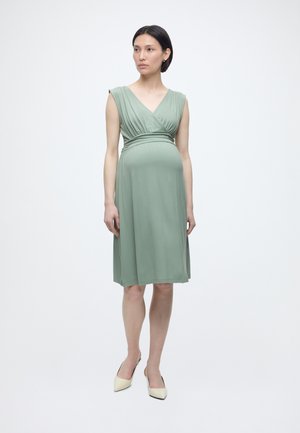 Terhes nő vékony színes, zsályazöld, térdig érő ruhában és krémszínű hegyes orrú magassarkú cipőben, egyszerű fehér háttér előtt áll.