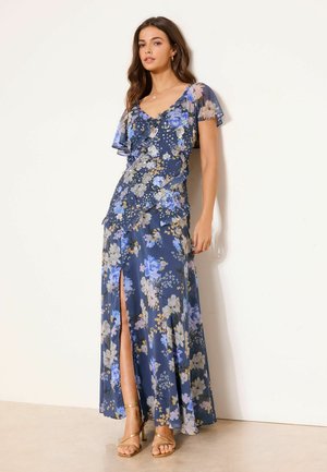 Mujer con vestido largo azul con estampado floral, mangas cortas con volantes y una abertura frontal, combinado con tacones dorados de tiras, de pie frente a una pared blanca.