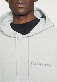 Sudadera gris hecha de una tela suave, con capucha ajustable y un logo bordado de "FILLING PIECES" en la parte frontal. Acabado texturizado visible.
