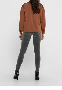 Braune Bluse mit langen Ärmeln und gepufften Schultern, kombiniert mit grauen Skinny-Jeans und schwarzen Stiefeletten mit hohen Absätzen. Einfaches Design ohne sichtbare Muster.