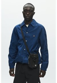 Veste légère bleu marine avec fermeture à boutons, manches longues et petit logo sur la poitrine. Associée à un sac noir et un pantalon foncé.