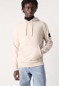 Felpa con cappuccio color crema, dotata di tasca frontale e orlo a costine. Presenta lacci regolabili e una patch nera con logo sulla manica sinistra.