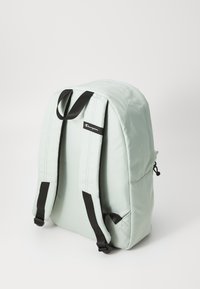 Sac à dos vert menthe avec un tissu lisse, des sangles noires réglables et une poche latérale. Présente un patch logo sur le devant.
