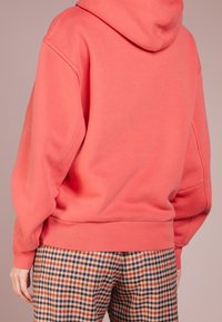 Koraalrode hoodie met een losse pasvorm en capuchon, gecombineerd met geruite broek in rood, blauw en geel. Zachte textuur en geribbelde boorden.