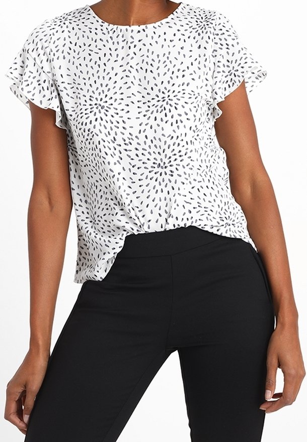 Femme portant un chemisier blanc à manches courtes avec un motif abstrait de points noirs et un pantalon noir taille haute, debout avec une main le long du corps.