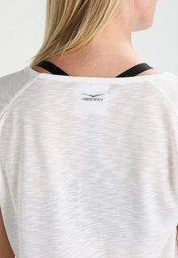 Camiseta atlética blanca con un acabado texturizado, presenta un logo negro "VENICE BEACH" en la parte posterior, con tirantes negros expuestos por debajo.