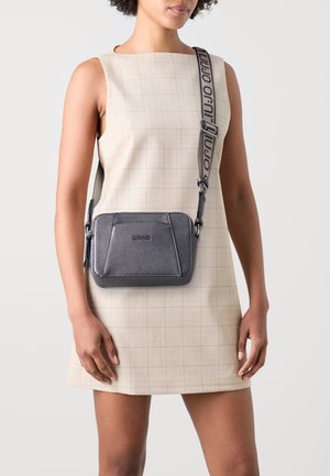 Borsa a tracolla grigia con tessuto testurizzato, forma rettangolare e cuciture a contrasto. Presenta un logo e una tracolla regolabile con testo.