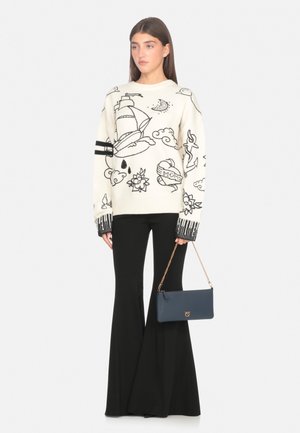 Donna che indossa un maglione bianco con disegni nautici e in stile tatuaggio neri, pantaloni neri a campana, mentre tiene una borsa blu con una catena dorata.