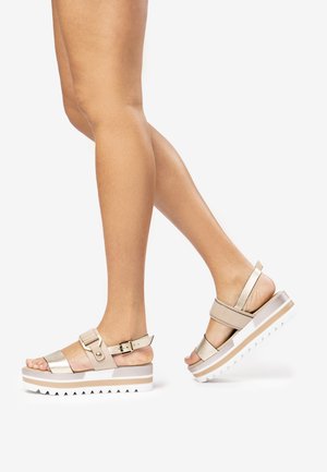Beige platform sandals with metallic straps, adjustable buckle, and textured white sole. Features an open toe design and wide ankle strap.