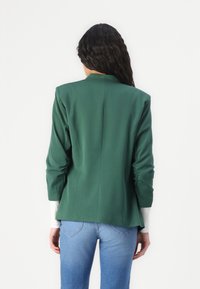 Zelený přizpůsobený blazer s vysokým límcem, dlouhými rukávy a peplum designem, kombinovaný se světle modrými džínami. Hladká látka, žádné viditelné vzory.