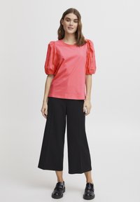 Tricou coral roz cu mâneci scurte din dantelă cu volane, asociat cu pantaloni negri cu croială largă și pantofi slip-on negri. Design simplu, casual.