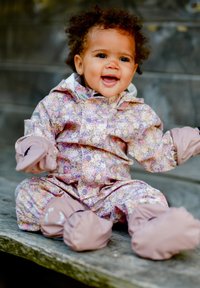 Kolorowy kombinezon przeciwdeszczowy z wzorem, z różowymi akcentami, wyposażony w kaptur, elastyczne mankiety i przymocowane ochraniacze na buty. Dziecko siedzi na drewnianej powierzchni.