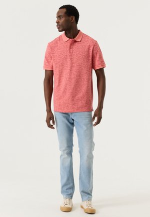 Man die een koraalroze polo met patroon en korte mouwen draagt, lichtblauwe spijkerbroek en beige sneakers, staand en zijwaarts kijkend tegen een effen achtergrond.