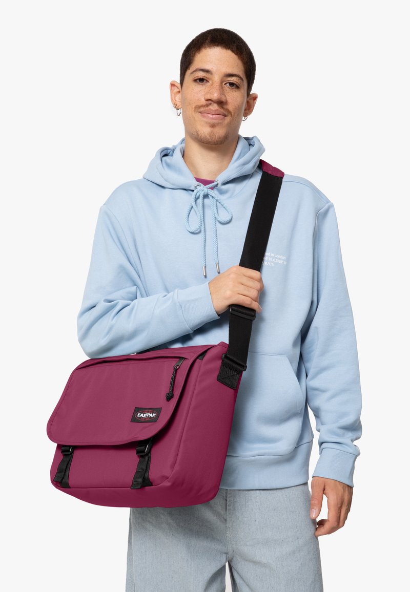 Purple messenger bag with flap closure, adjustable strap, and front zip pocket. Made from durable fabric, features logo patch and smooth texture.