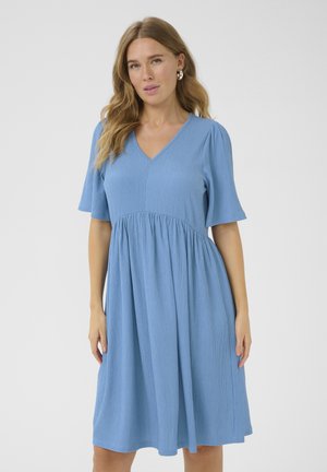 Femme aux cheveux ondulés portant une robe midi bleu clair à manches courtes, col en V, taille froncée, debout devant un fond uni.