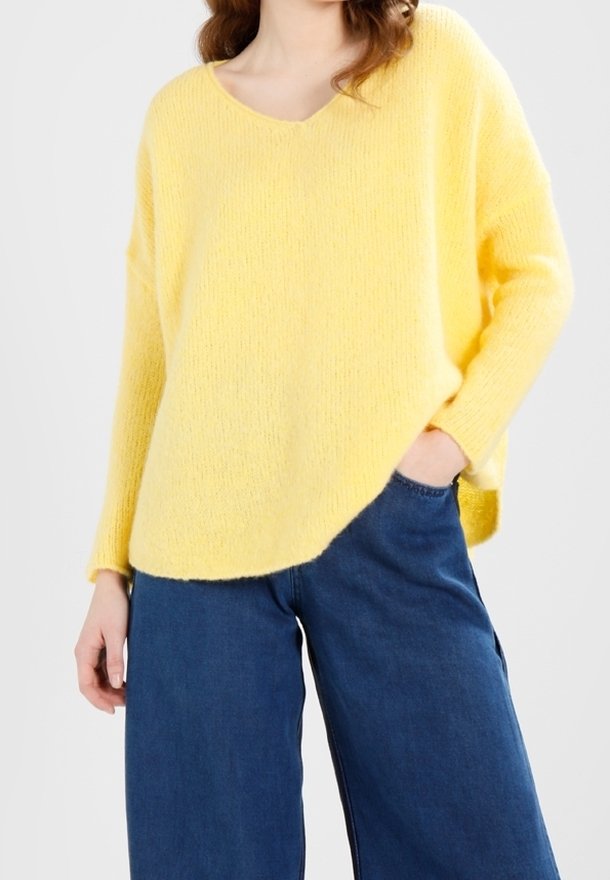 Femme portant un pull jaune ample à col en V avec des manches longues et un jean bleu à jambes larges, une main dans la poche, debout devant un fond blanc.