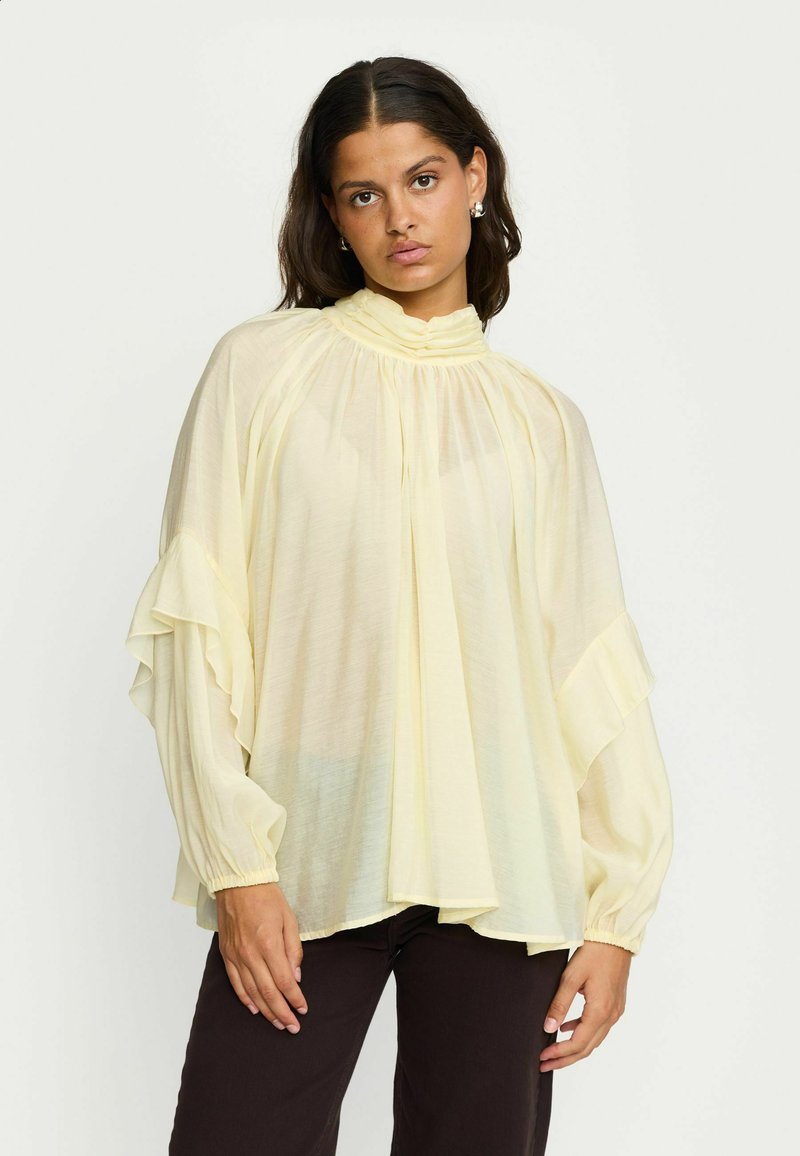 Blouse jaune clair en tissu semi-transparent. Présente un col montant, des détails plissés et des manches volantes. Design doux et fluide.